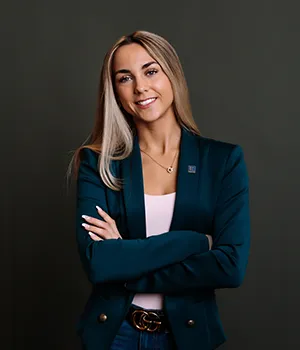 Woman with long blonde hair in a teal blazer stands smiling with arms crossed.