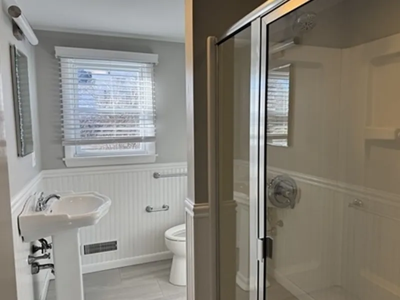 A bathroom with a glass shower, toilet, sink, and a window with blinds.