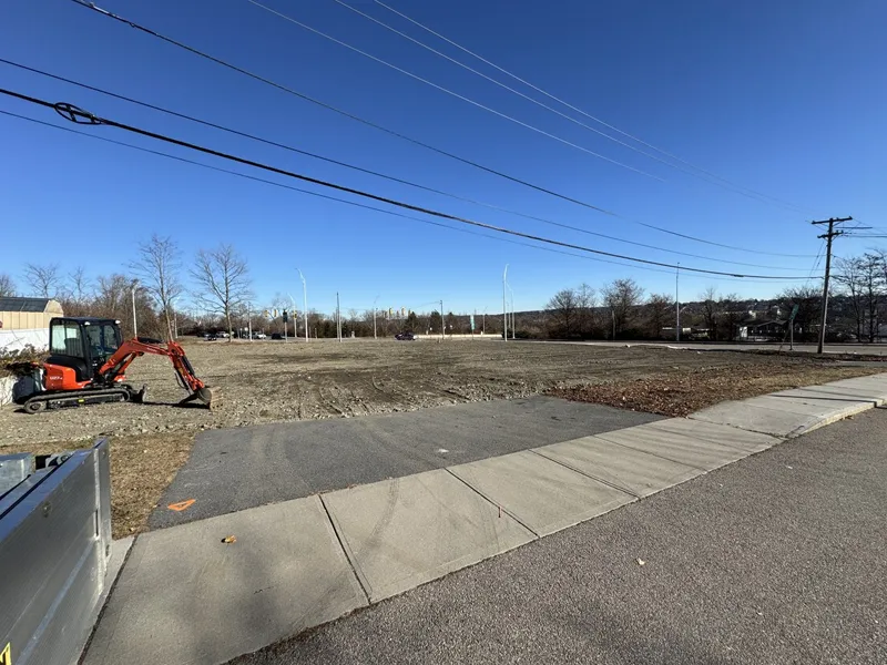 An empty lot with a small excavator beside a sidewalk under a clear blue sky.