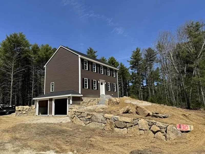 Two-story brown house with garage, stone retaining wall, and dirt yard surrounded by trees.