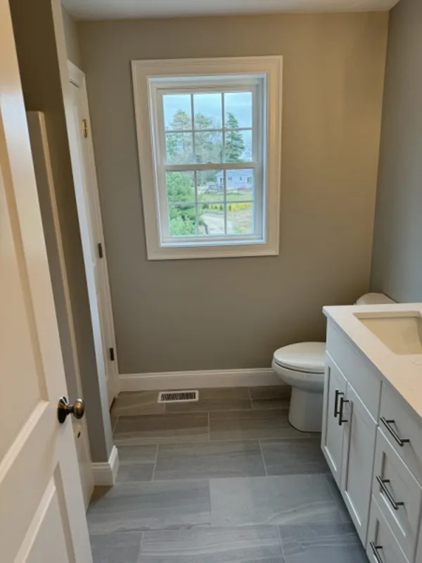 Small bathroom with gray tile floor, white vanity, toilet, and window showing greenery outside.