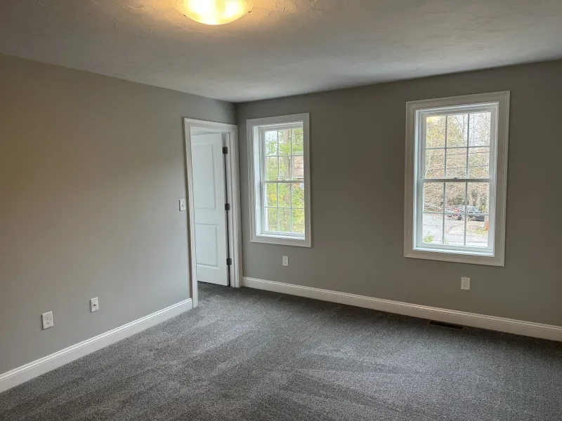 Empty room with gray walls, carpeted floor, two windows, and a closed white door.