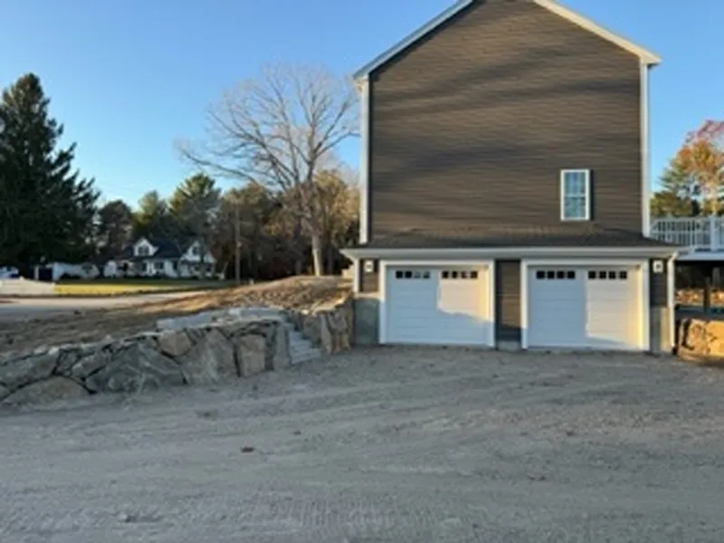 Two-car garage with gravel driveway and stone retaining wall, trees and houses in background.