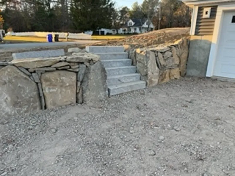Stone steps set between large rocks lead from a gravel driveway to a higher yard level.