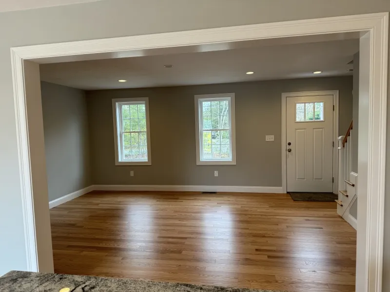 Empty room with light wood floors, gray walls, two windows, and a white front door.