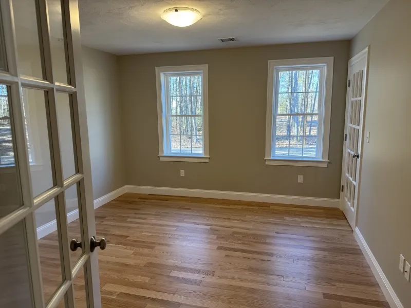 Empty room with wood floors, beige walls, two windows, and a glass door.