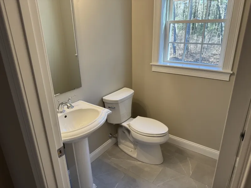 Small bathroom with a pedestal sink, toilet, mirror, window, and beige walls.