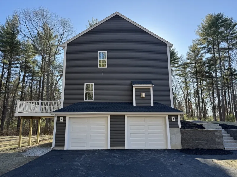 Three-story gray house with double garage, deck, and surrounding trees on a sunny day.