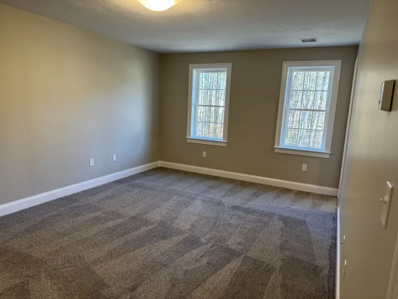 Empty room with beige walls, two windows, and gray carpet flooring.