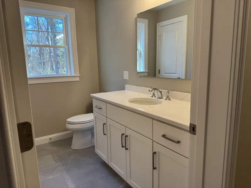 Small bathroom with a white vanity, sink, mirror, toilet, and window letting in natural light.
