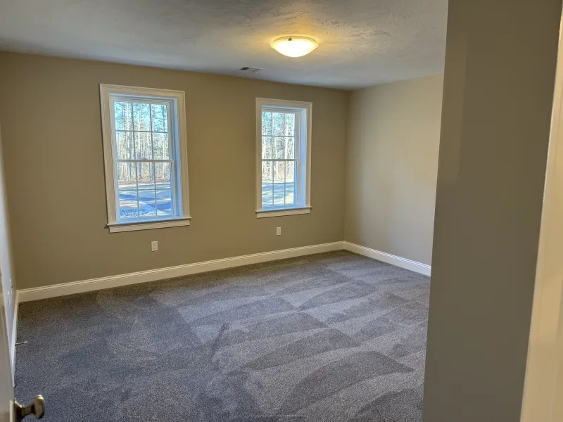 Empty room with beige walls, gray carpet, and two windows letting in natural light.