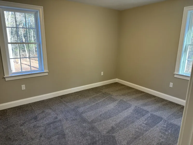Empty room with beige walls, gray carpet, and two windows letting in natural light.