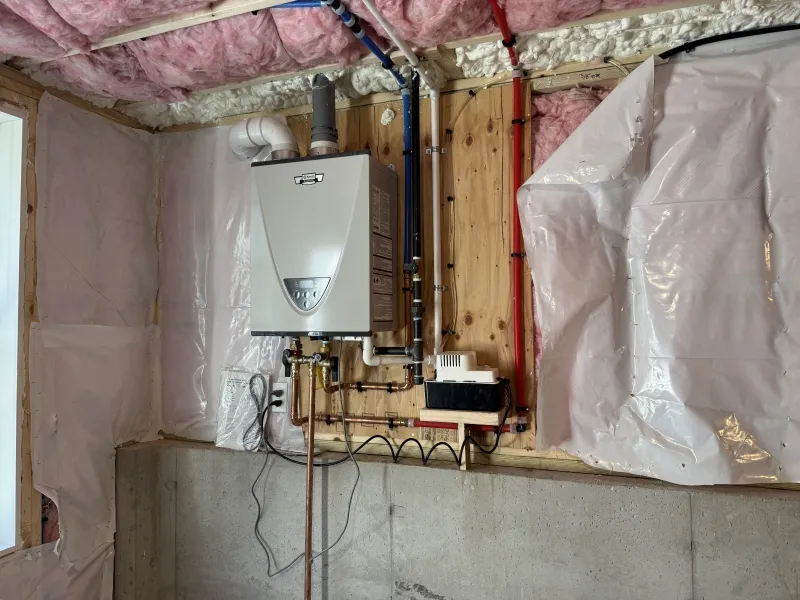 Tankless water heater mounted on a basement wall, surrounded by pipes and insulation.
