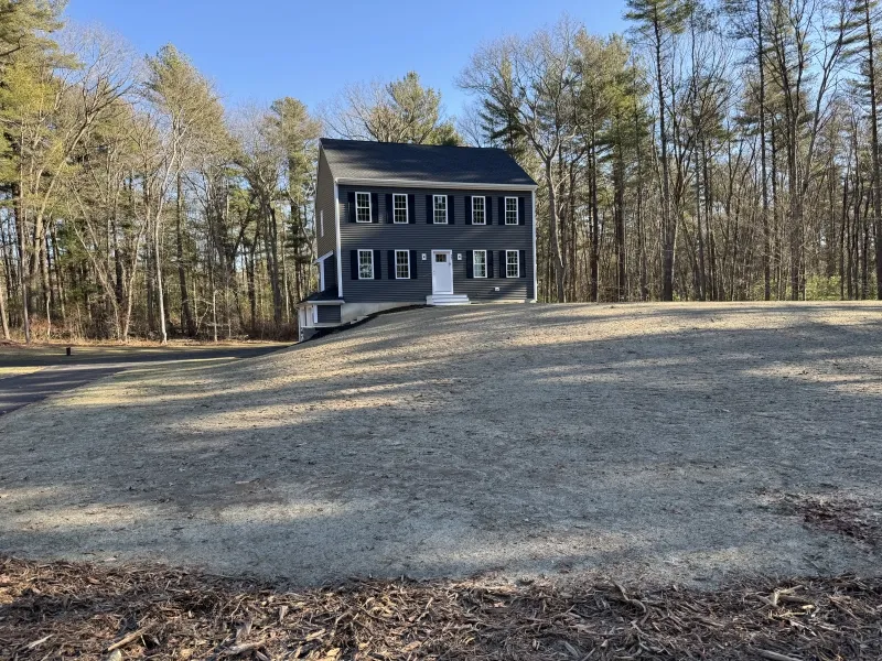 A two-story dark gray house sits on a large, sloped lawn with trees in the background.