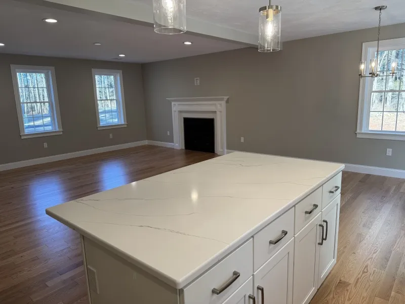 Kitchen island with white countertop overlooking empty living room with fireplace and windows.