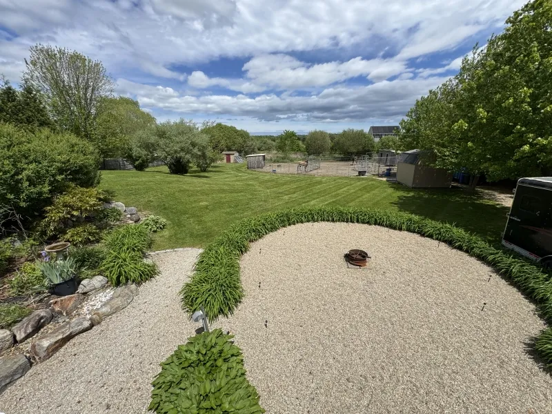 Gravel patio with fire pit, surrounded by green lawn, trees, and a partly cloudy sky.