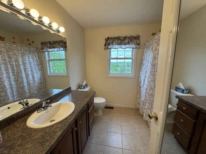 Bathroom with double sinks, large mirror, toilet, window, and patterned shower curtain.