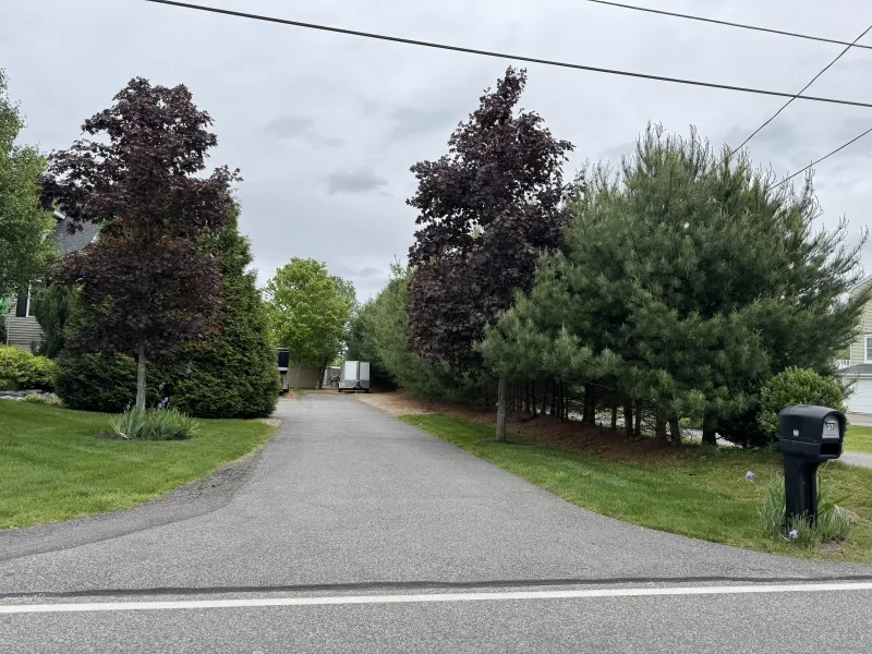 A paved driveway lined with trees leads to a white trailer under a cloudy sky.