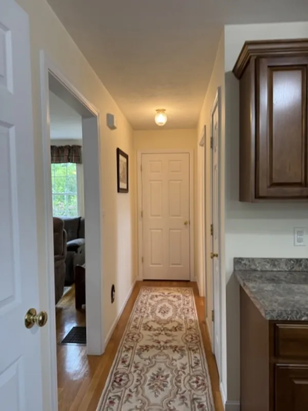 A hallway with a patterned runner, light walls, and a door at the end, near a kitchen.