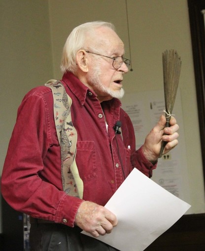 Older man in red shirt holds a fan-shaped object and paper while speaking indoors.
